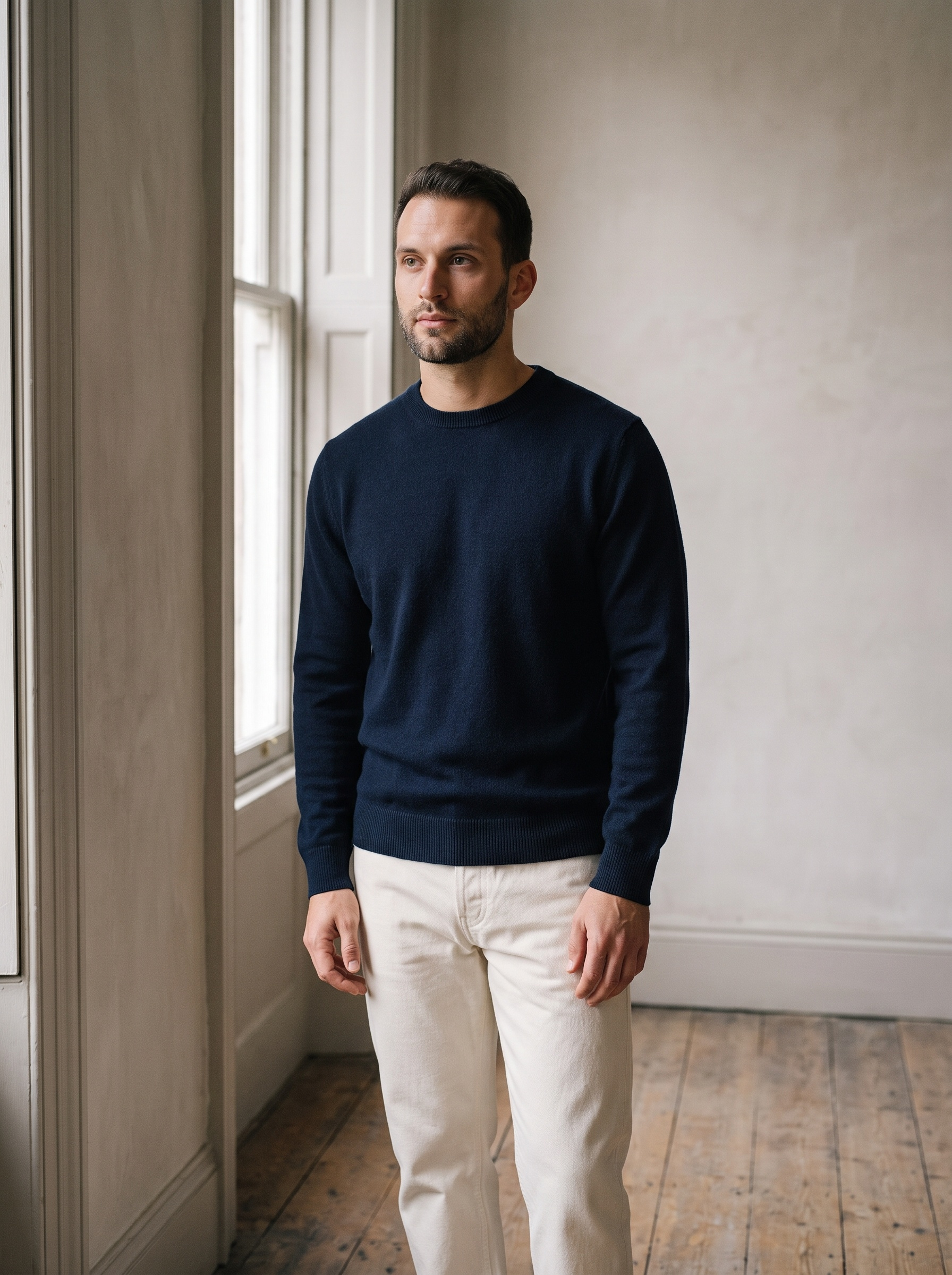 A person in a navy knit and cream trousers standing by a tall window in daylight